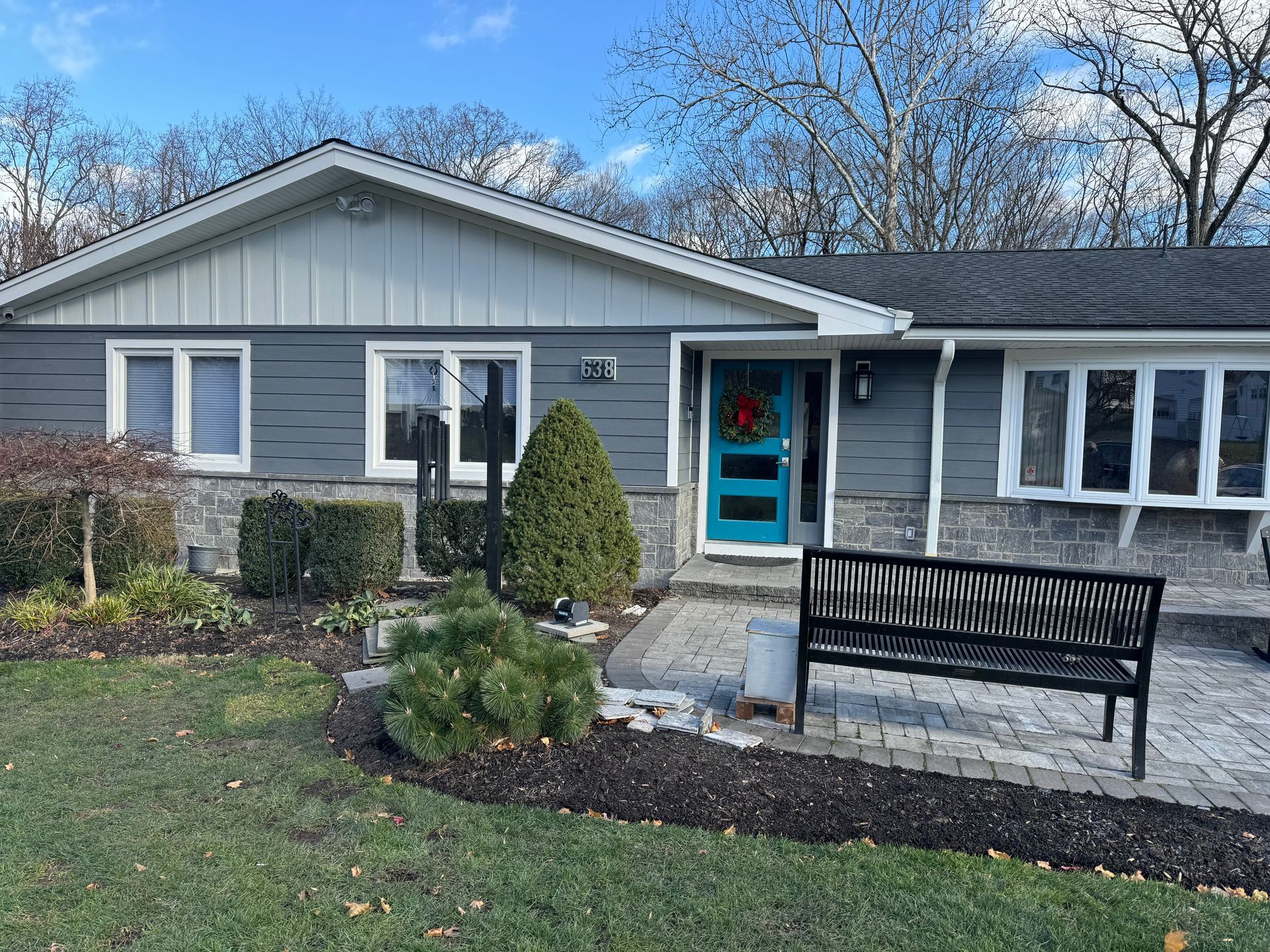 Single-story gray house with teal front door, shrubs, and a bench by the walkway under a blue sky