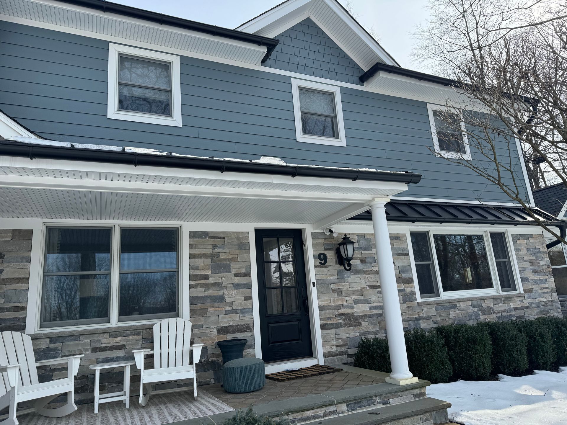 Blue-gray house with stone facade, black door, white porch columns, and snow on the ground