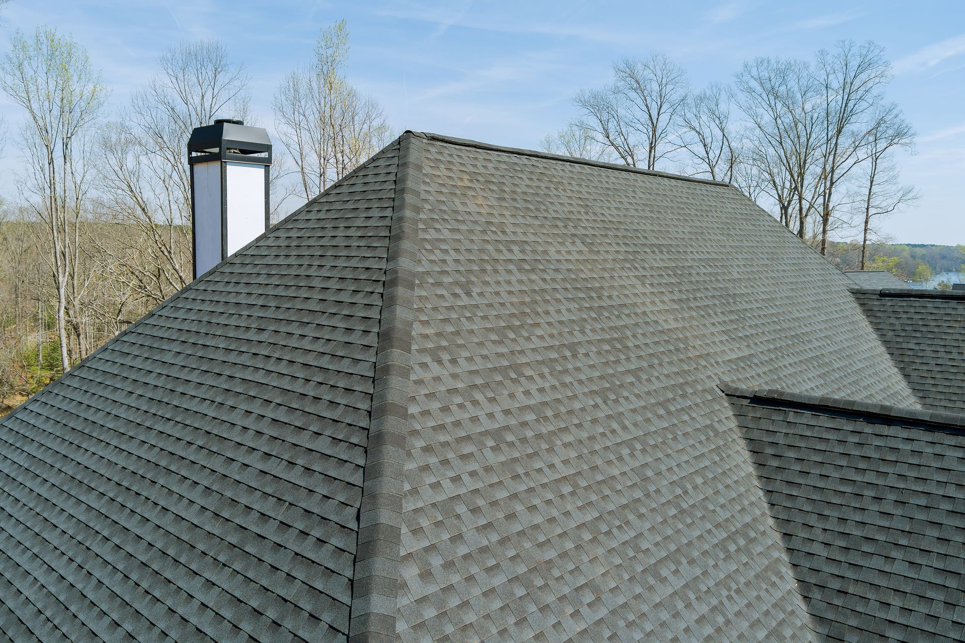 Gray shingled roof with chimney against leafless trees and blue sky