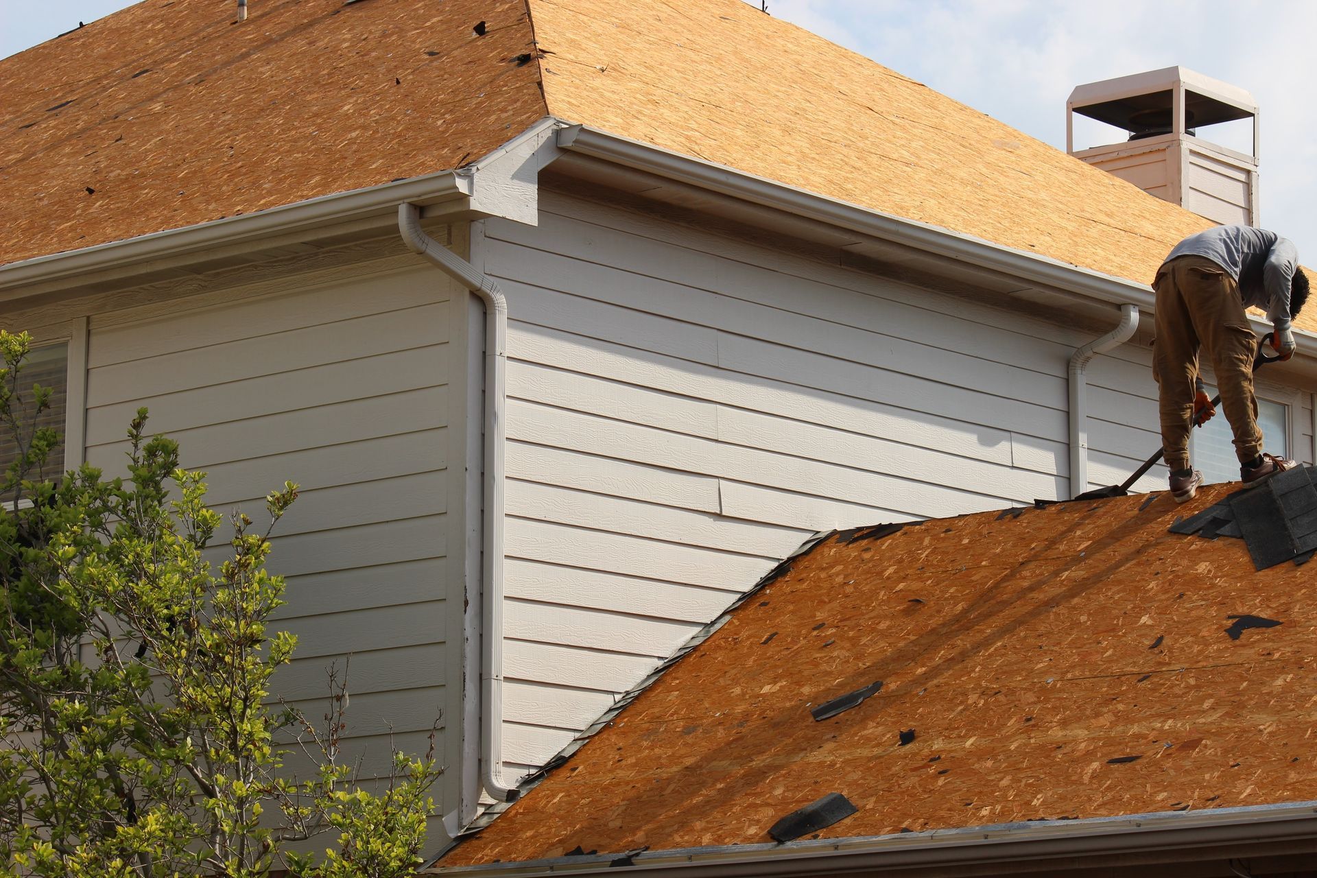 Worker on an orange roof repairing shingles beside a white house exterior.