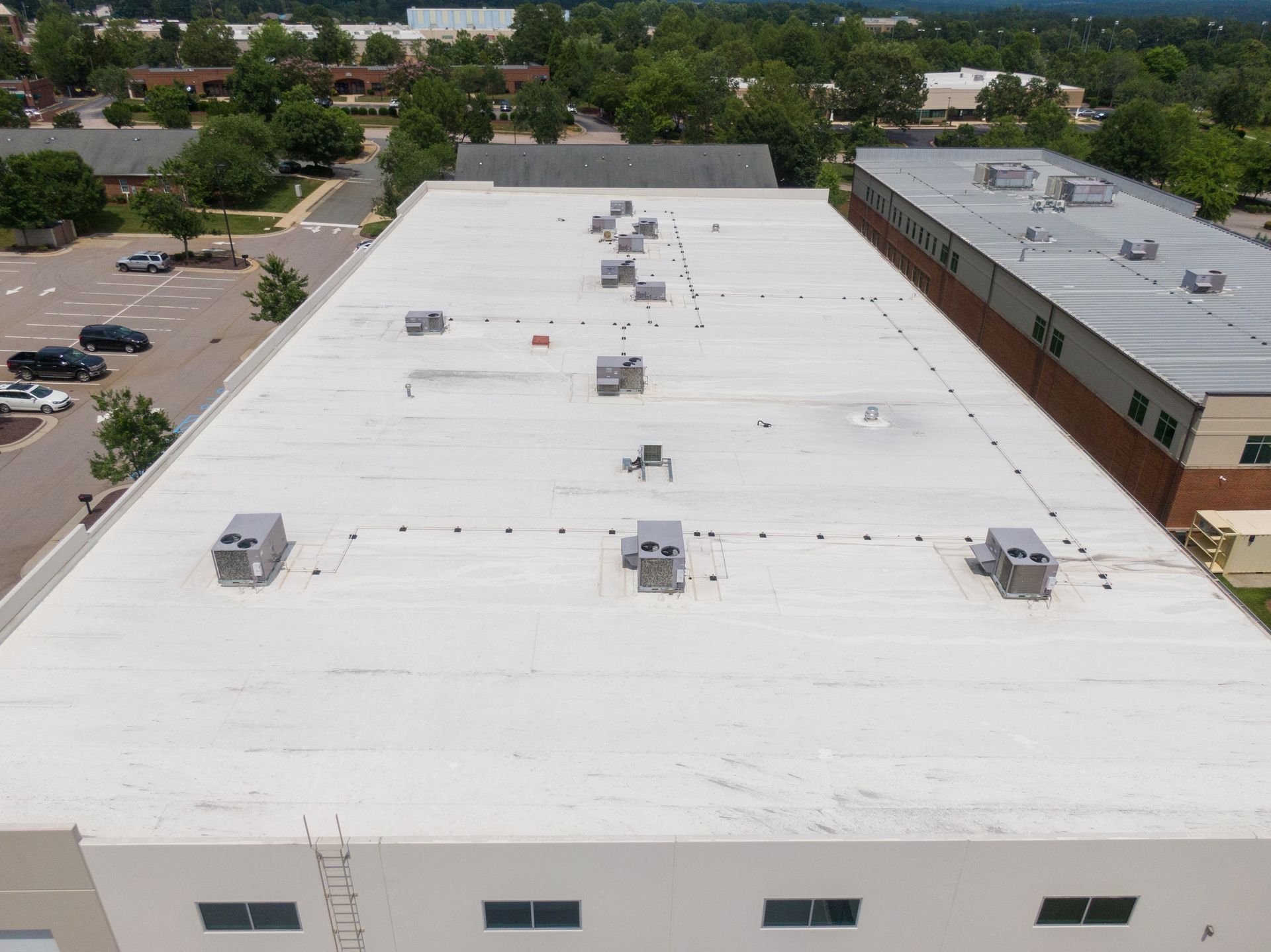 Aerial view of a large white-roofed industrial building with rooftop units and surrounding trees and parking lots