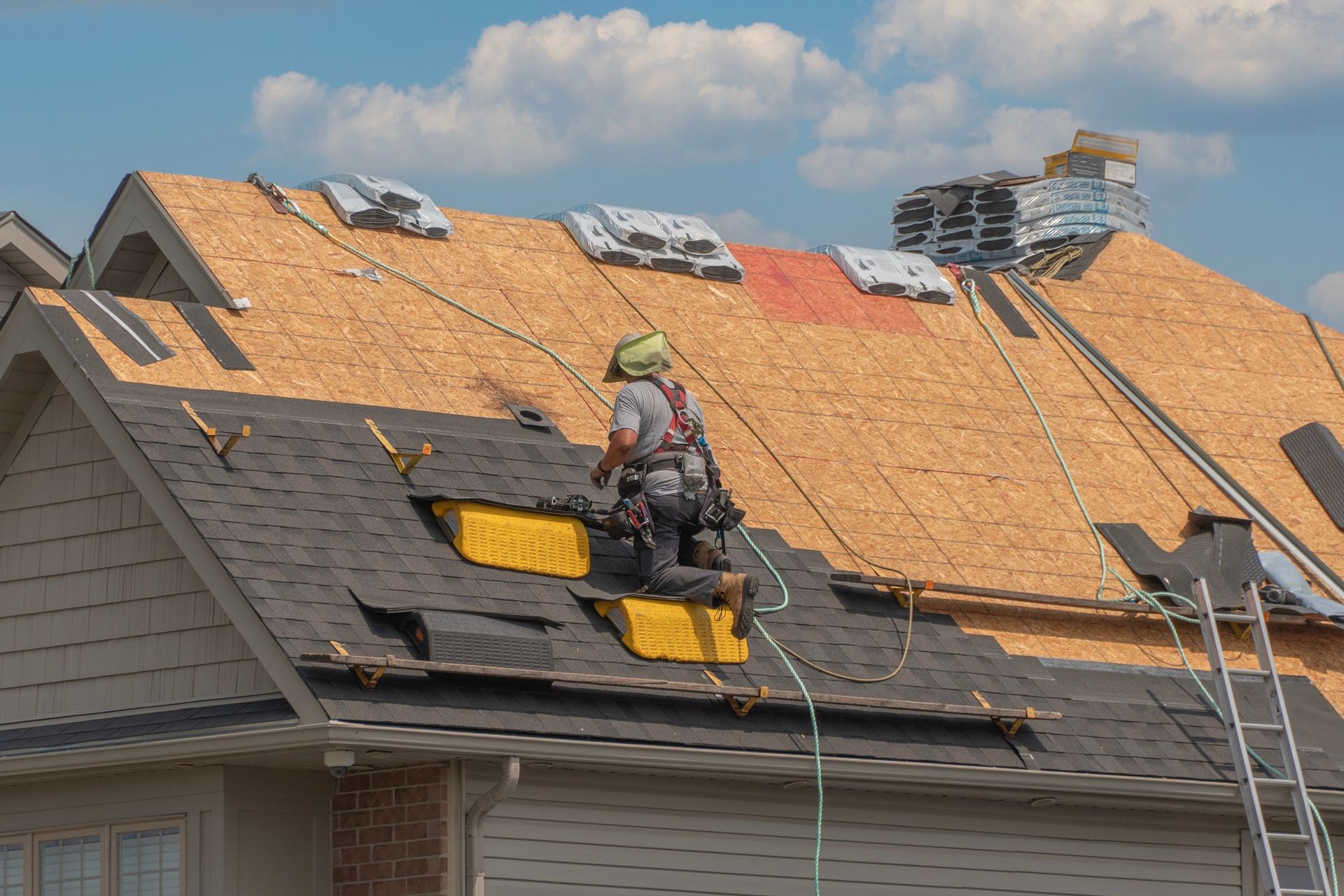 Roofer in safety gear working on a house roof, replacing shingles. Ladders and materials visible.