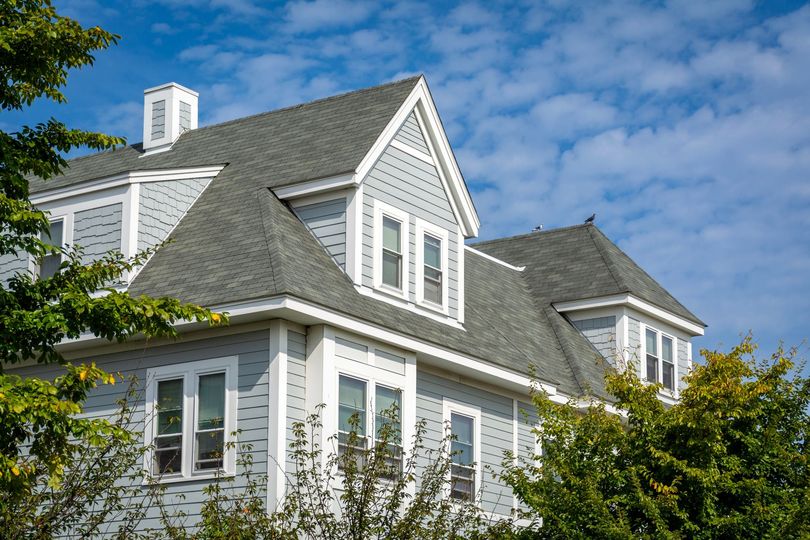 Two-story light blue house with gray roof, white trim, and multiple windows; trees and blue sky.