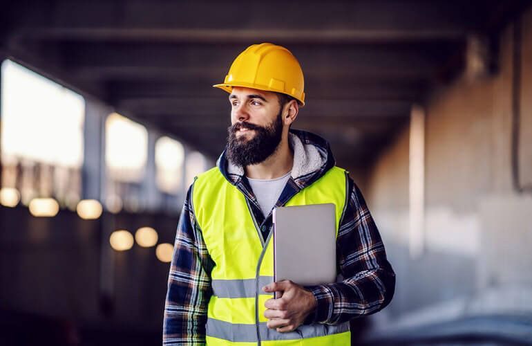 Construction worker in blue hard hat and safety vest, standing with arms crossed at a building site.