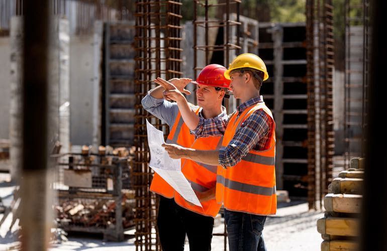 Construction worker in blue hard hat and safety vest, standing with arms crossed at a building site.