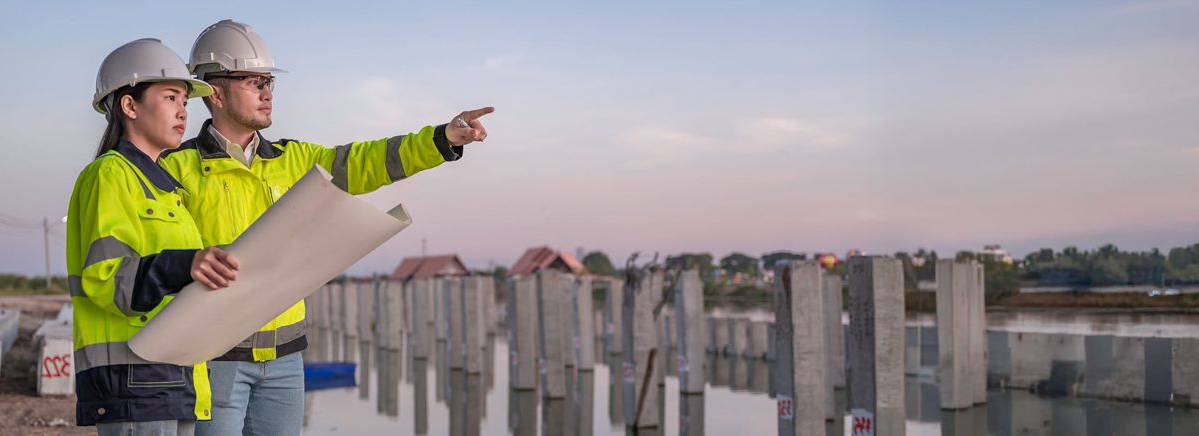 Two construction workers in hard hats review plans atop a building site at sunset