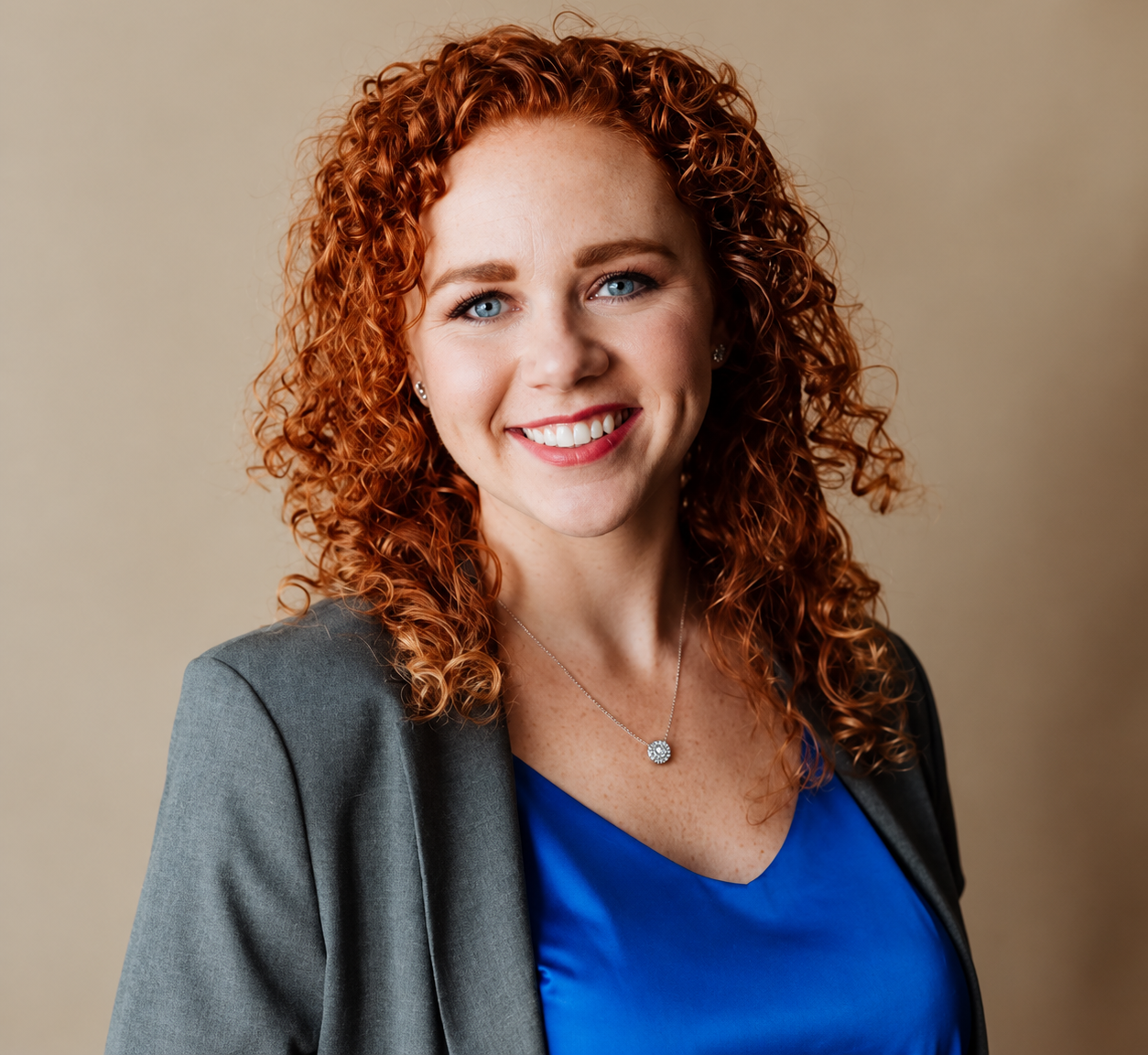 Headshot of a smiling person with curly auburn hair, wearing a grey blazer, against a neutral background.