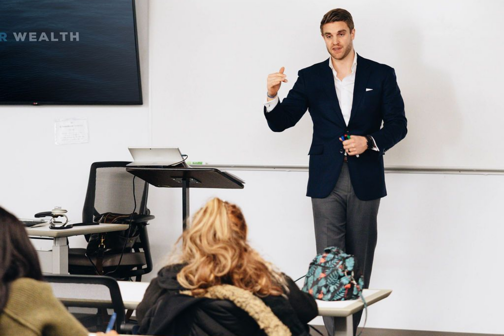 A speaker in a dark blazer gestures while presenting to an audience in a bright room with a monitor and podium.