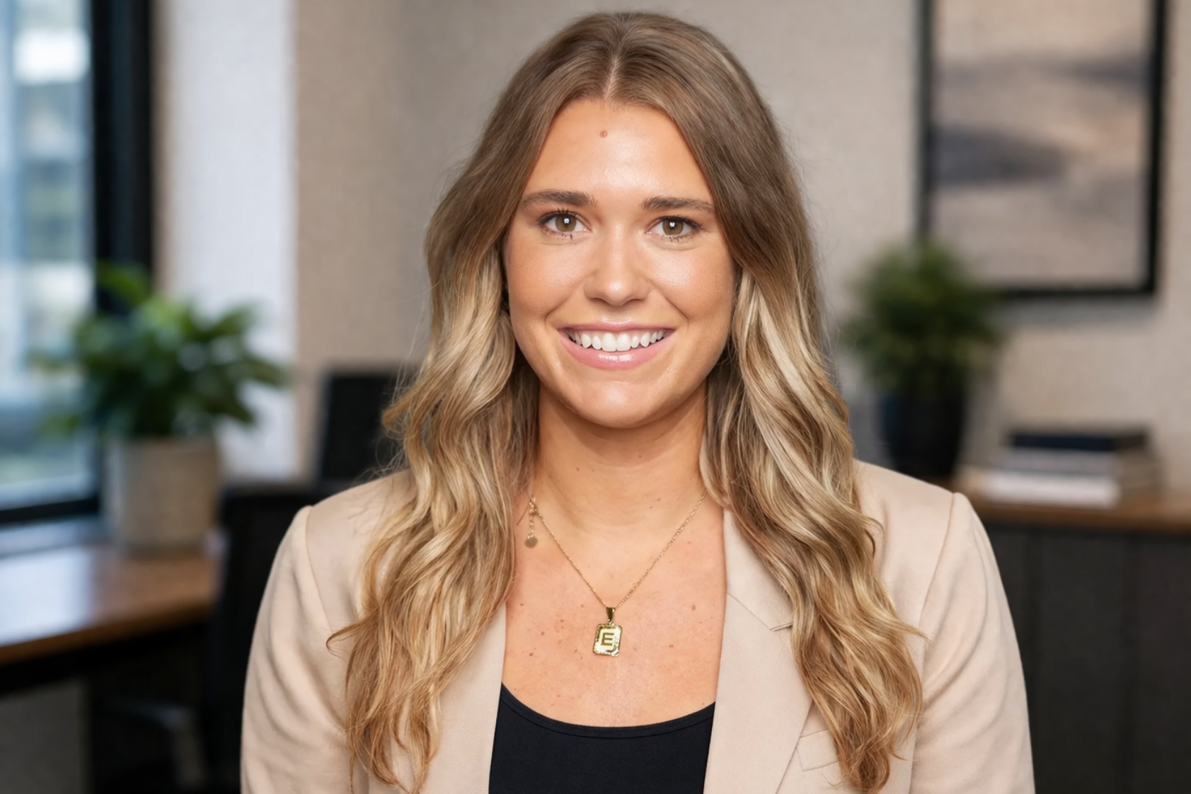 A professional portrait of a person with long blonde hair, wearing a tan blazer and gold necklace, posing in an office.