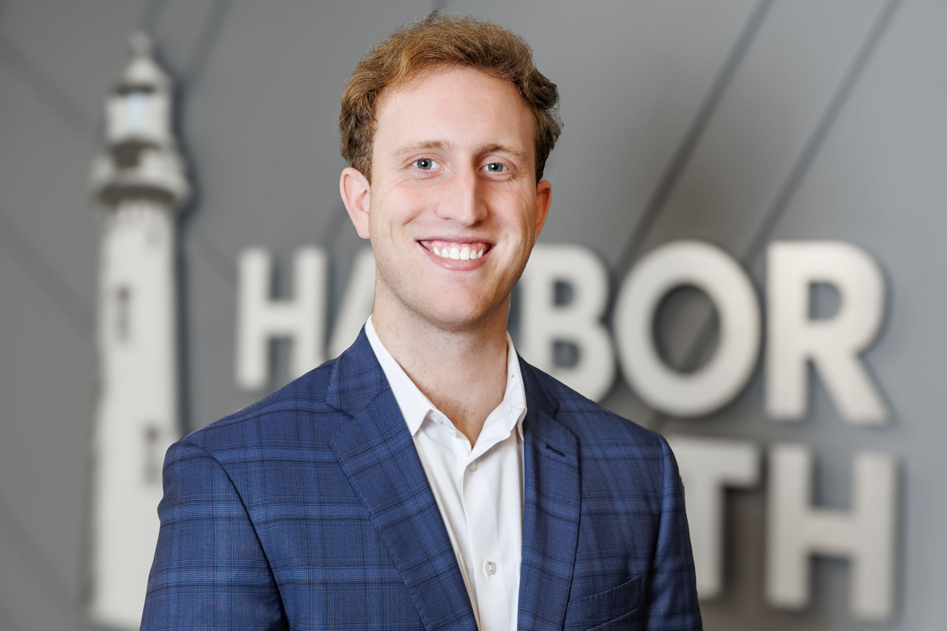 A person smiling in a blue plaid blazer and white shirt, standing in front of a gray wall with a lighthouse logo.