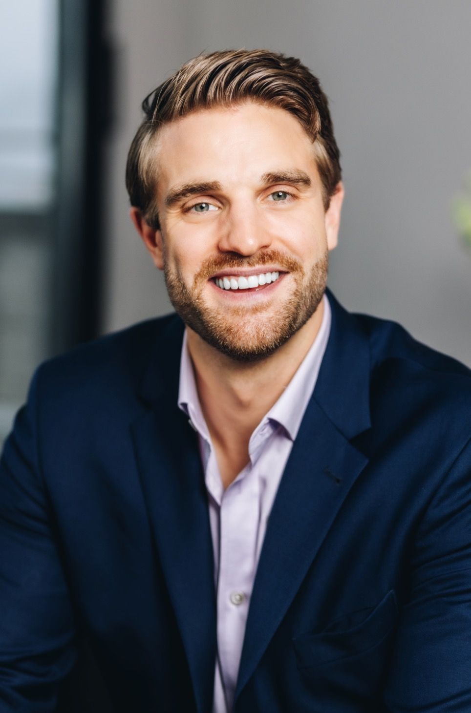 Smiling man in a navy blazer and light shirt, posed against a softly blurred gray background