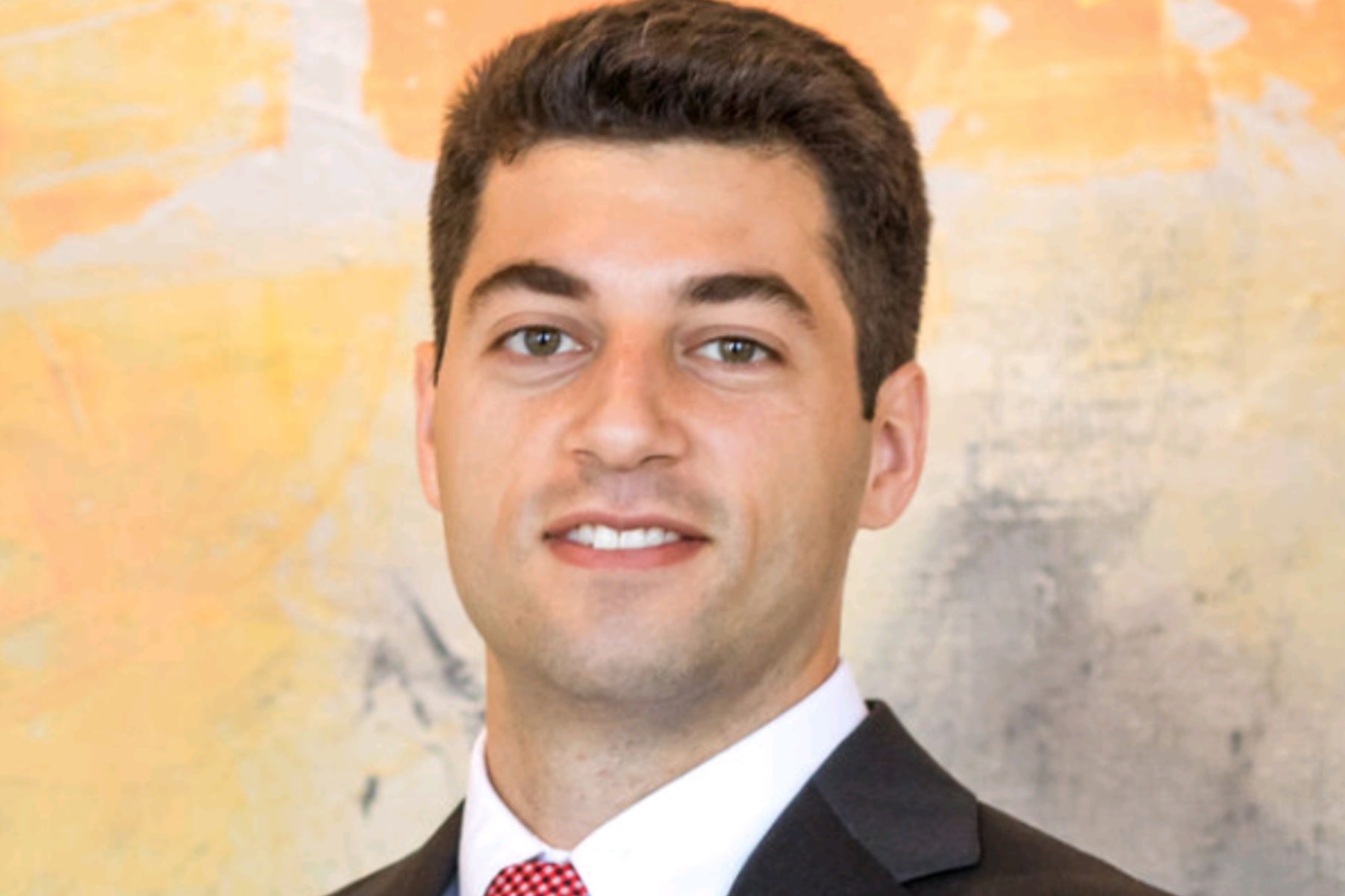 Headshot of a smiling person wearing a dark suit, white shirt, and patterned red tie against an abstract painted backdrop.
