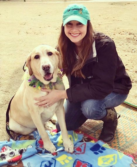 Woman sitting with a brown dog, both smiling. The dog is panting, and the woman is wearing a yellow shirt.