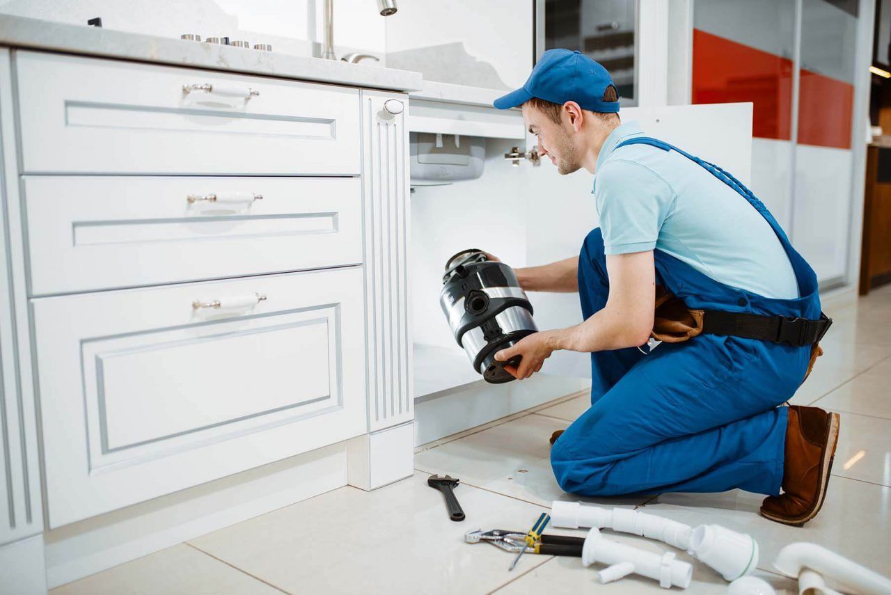 Plumber in blue uniform repairing a white kitchen cabinet sink with tools on the floor