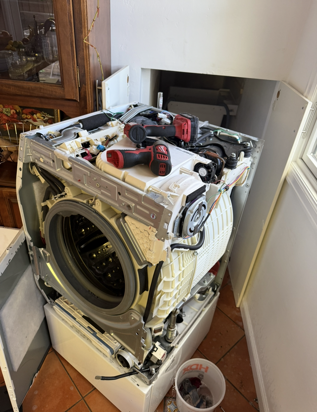 Damaged front-loading washer with tools on top, sitting in a laundry area with an open wall behind it