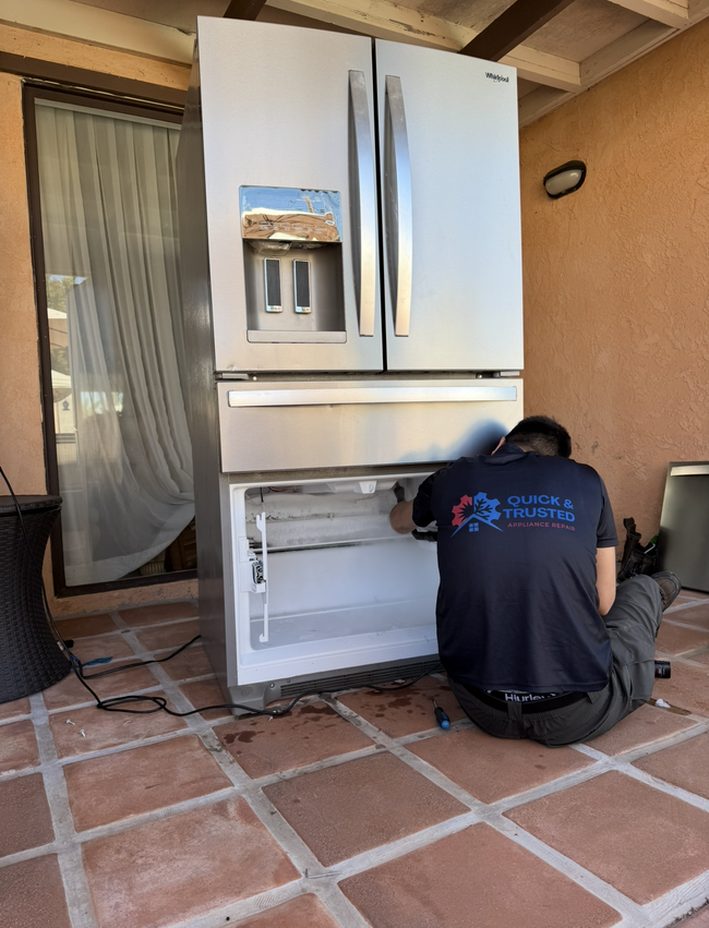 Technician kneeling beside a white refrigerator, working on the open lower freezer door on a tiled patio.