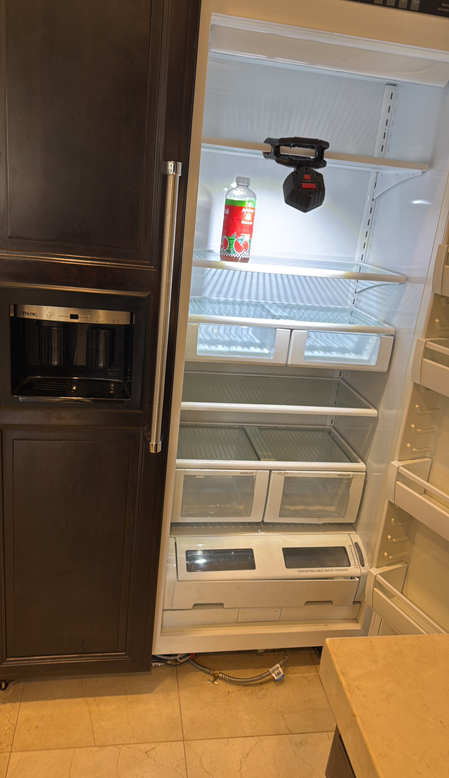 Empty built-in refrigerator beside dark wood cabinets, with shelves and a beverage bottle inside.