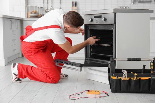 Repairman kneeling by a built-in oven, using a screwdriver with tools on the floor in a bright kitchen