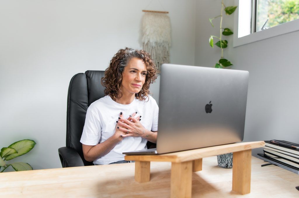 Woman with curly gray hair smiling, hands on her chest, video conferencing on a laptop in a home office.