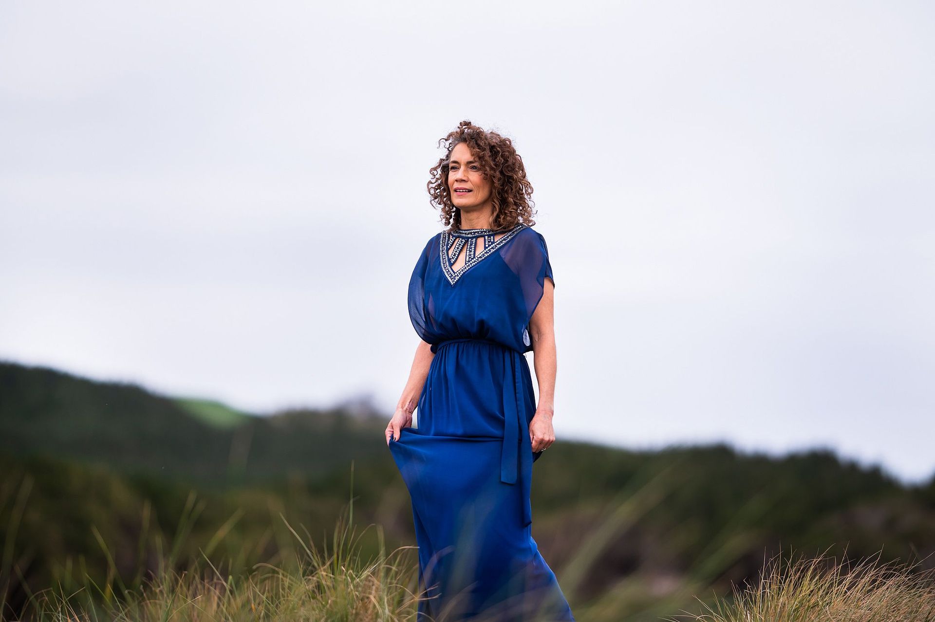 Woman with curly hair wearing a long blue dress, standing in tall grass on a cloudy day.