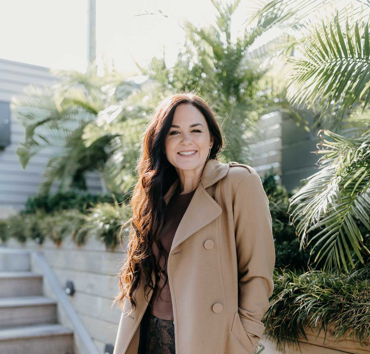 Woman with long brown hair smiles, wearing a tan coat, standing outdoors in front of greenery.