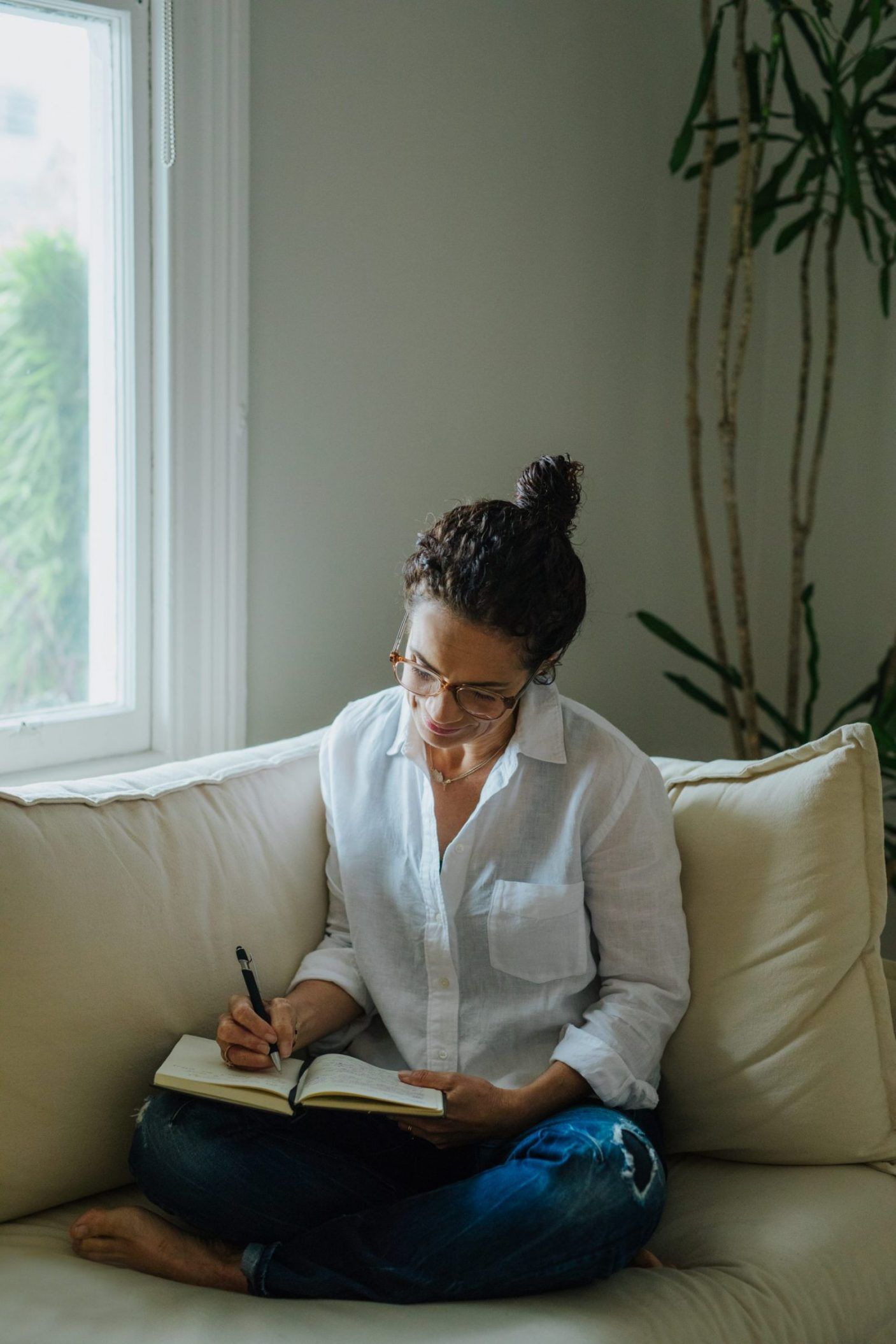 Tracy Manu with glasses writing in a notebook while sitting cross-legged on a couch by a window.