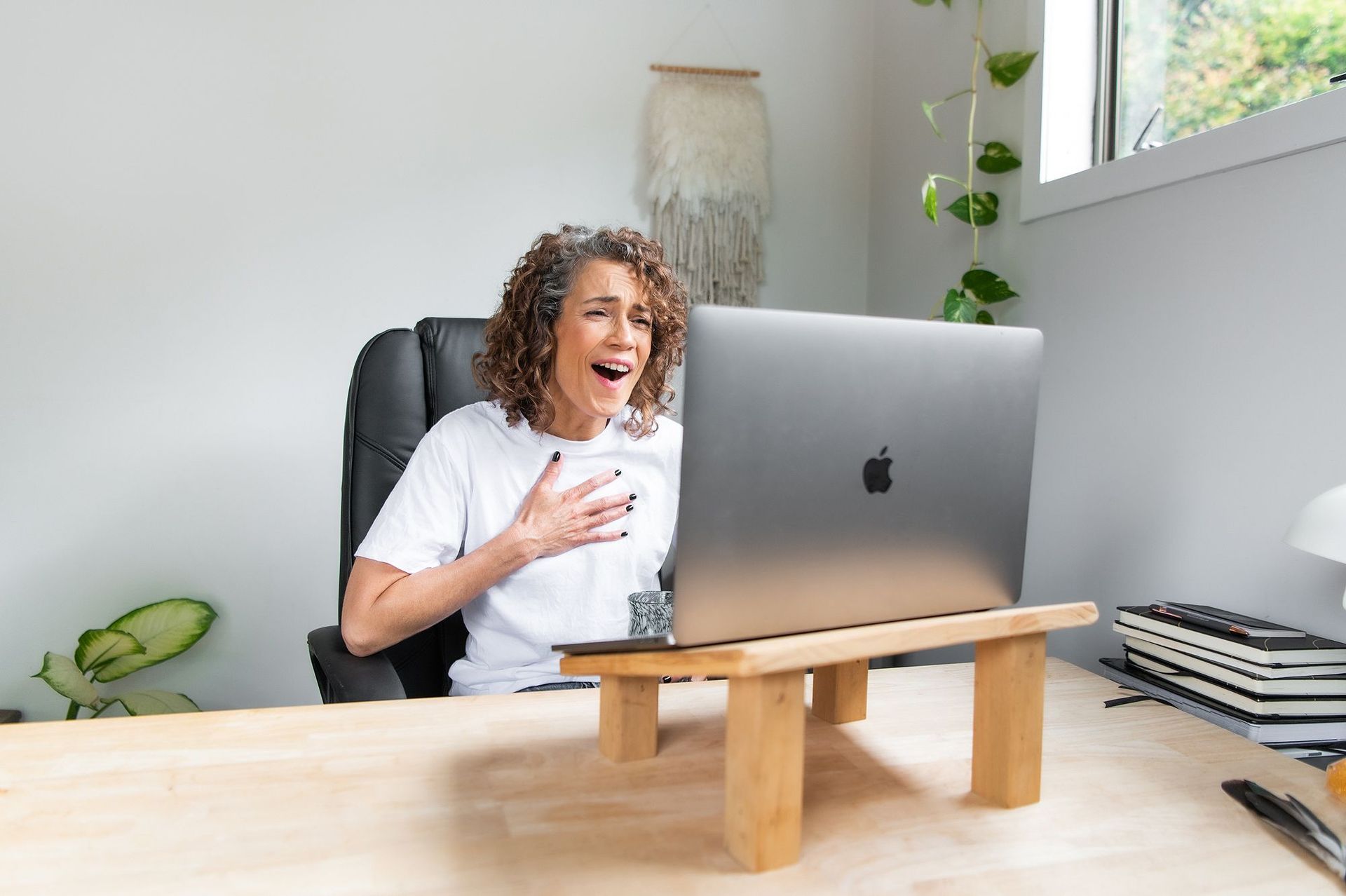 Tracy Manu with curly hair reacts with surprise while looking at a laptop on a wooden stand, at a desk in a bright room.