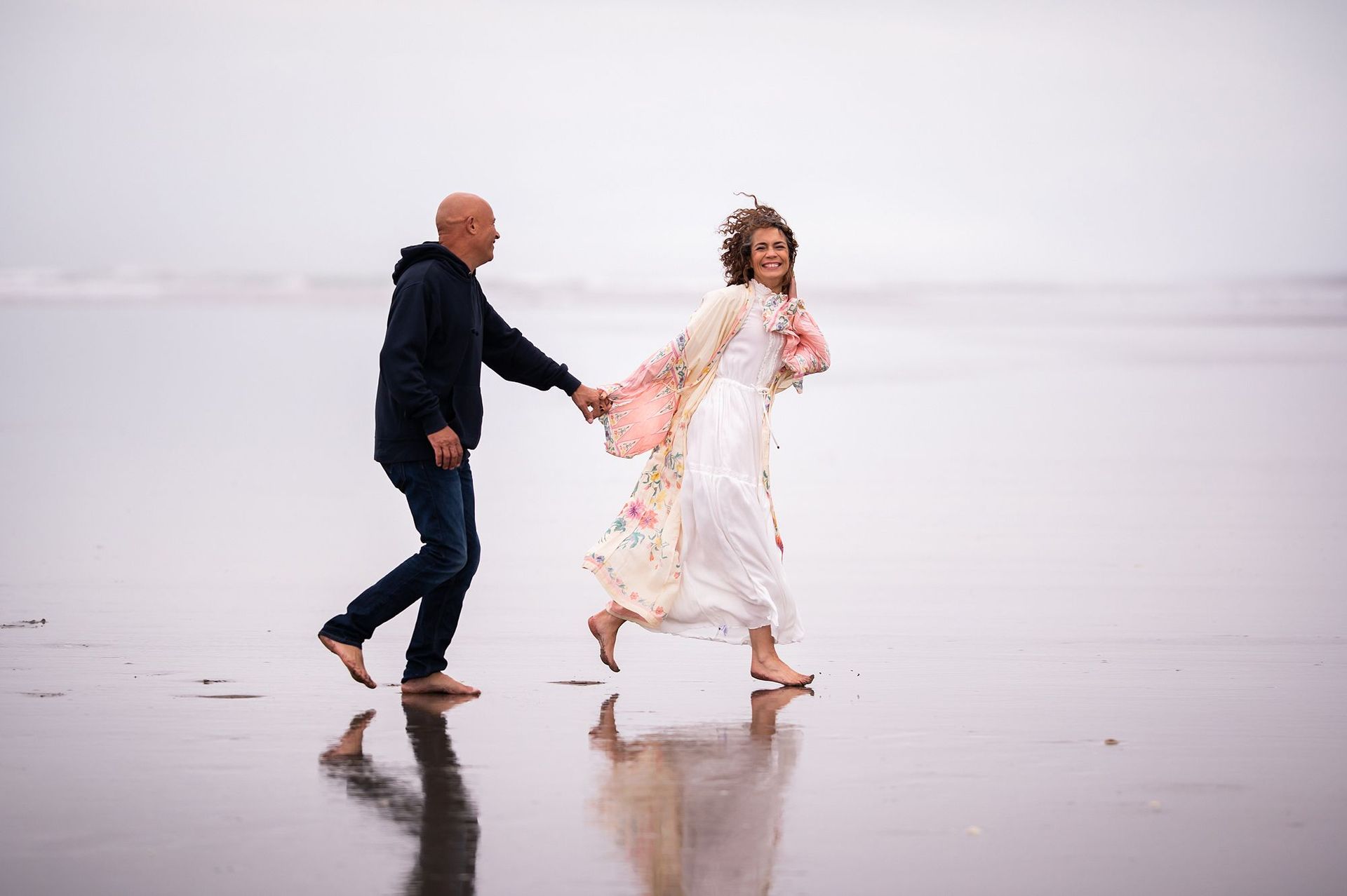 A barefoot couple running on a wet beach, the man in a hoodie and jeans, the woman in a floral dress, holding hands, smiling.