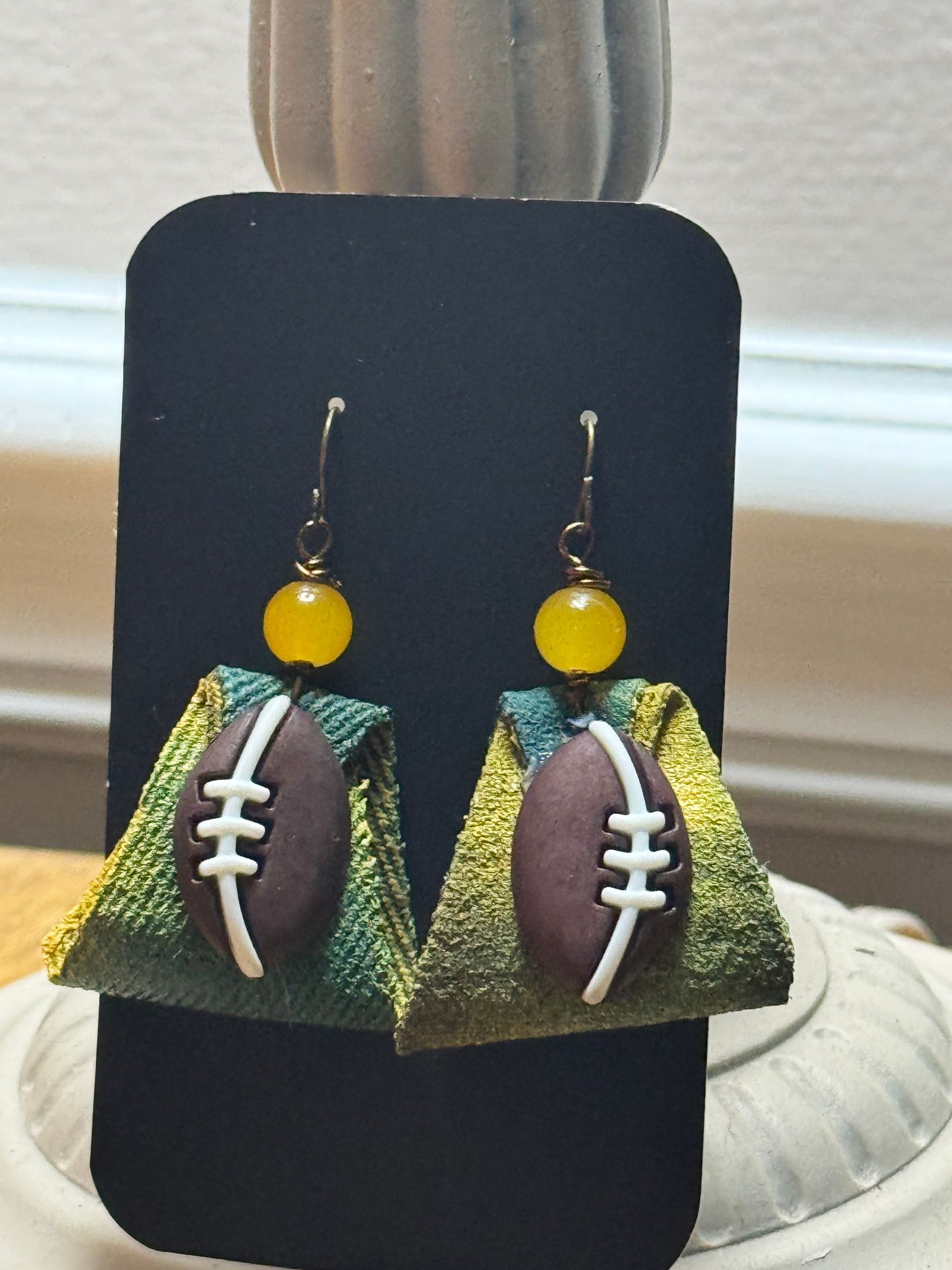 Football-shaped earrings with yellow beads on a black display card, with white, green, and brown detailing