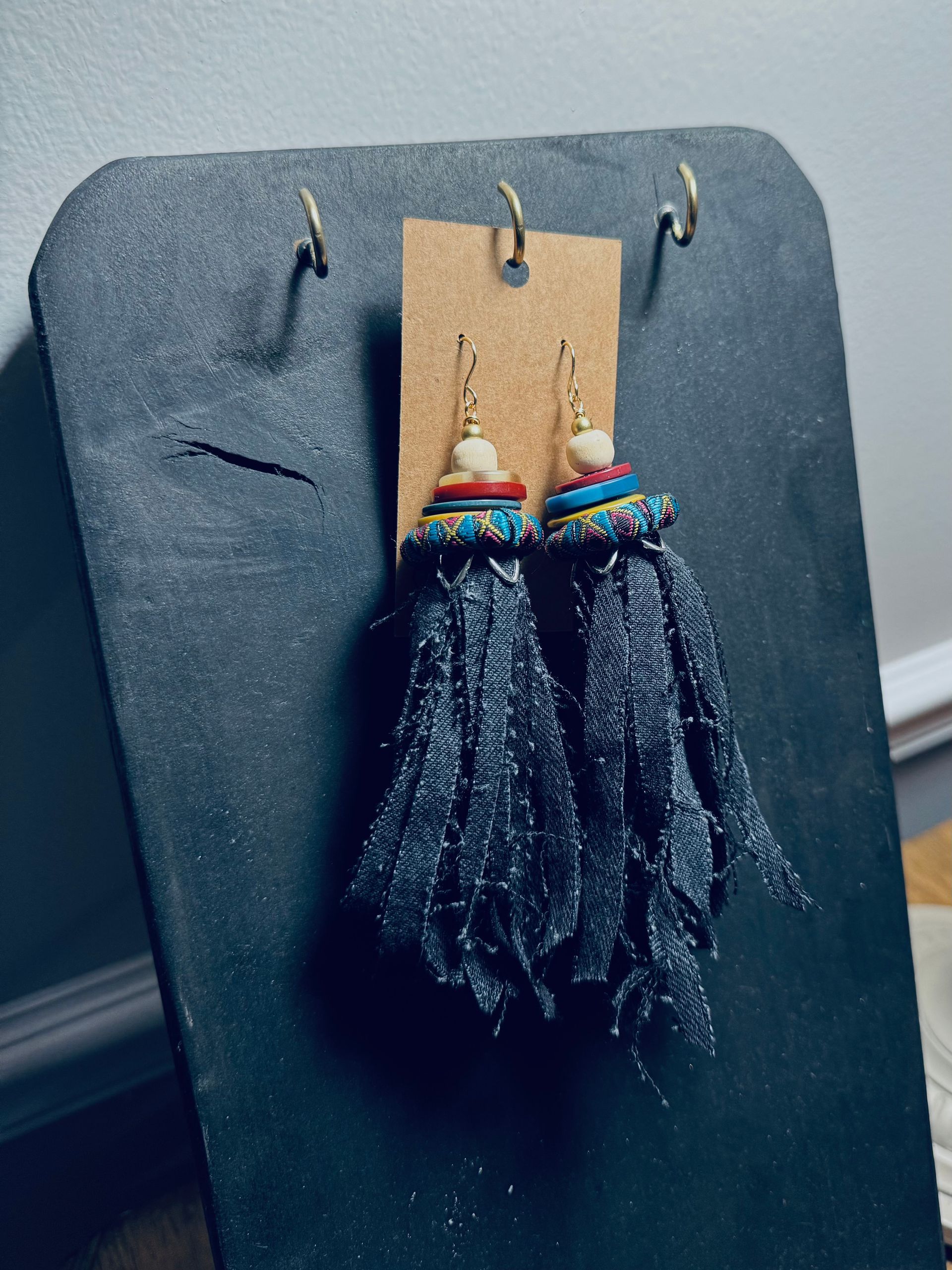 Black felt earrings on a cork card with multicolored beads and black fringe, hanging on a dark display board