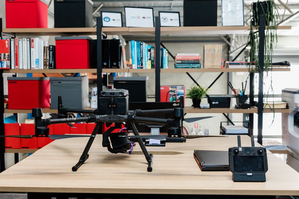 Drone on a desk with remote and laptop, in front of a shelf filled with office supplies.