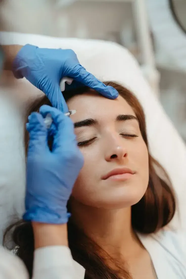 Gloved hands apply cosmetic treatment to a woman’s forehead with a syringe in a clinic