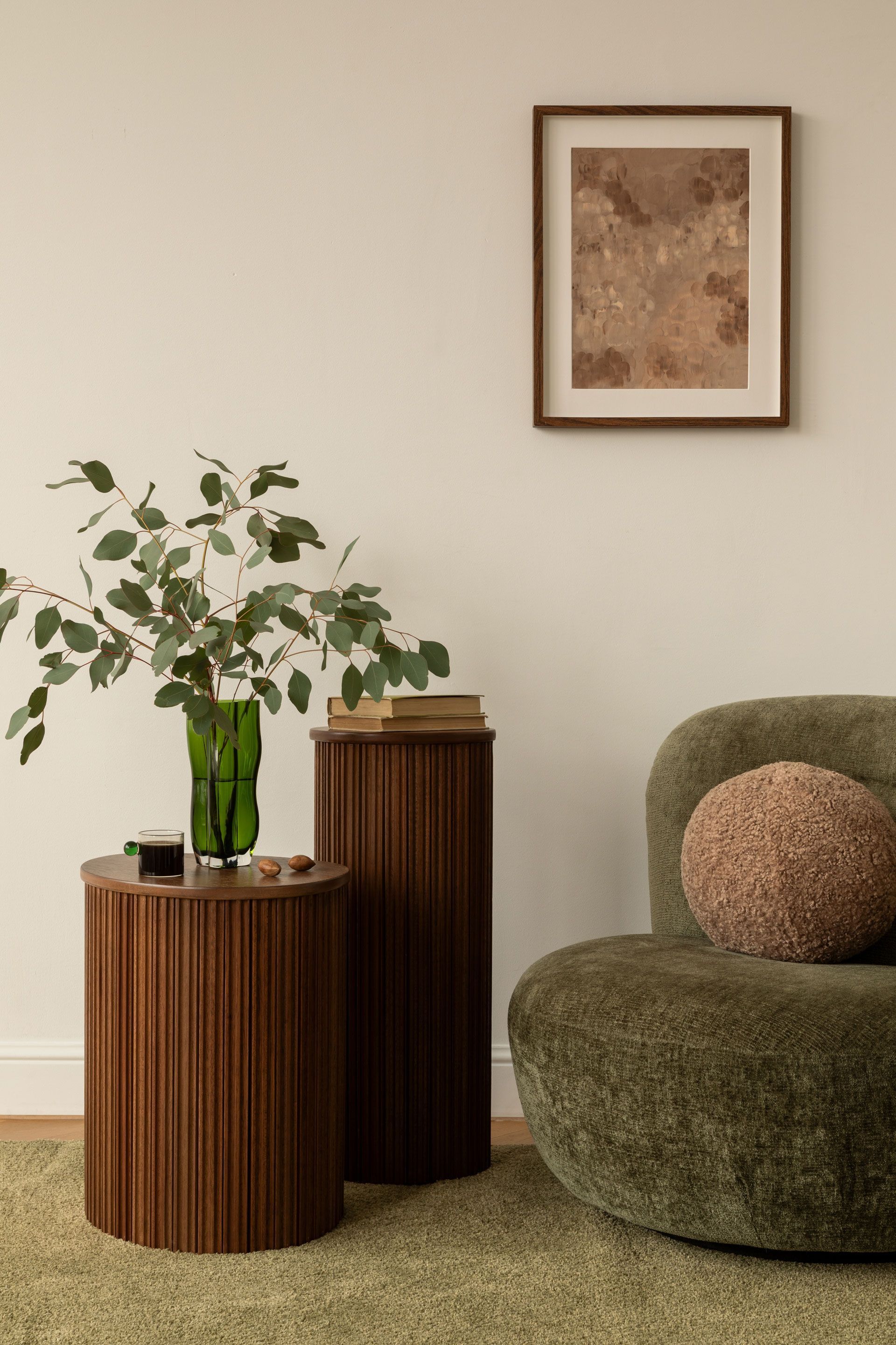 Cozy living room corner with green chair, wooden side tables, a vase of branches, and framed wall art