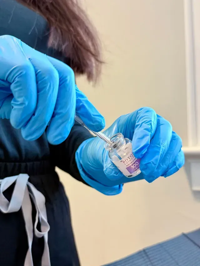 Blue-gloved hands hold a small clear vial and dropper in a clinical setting