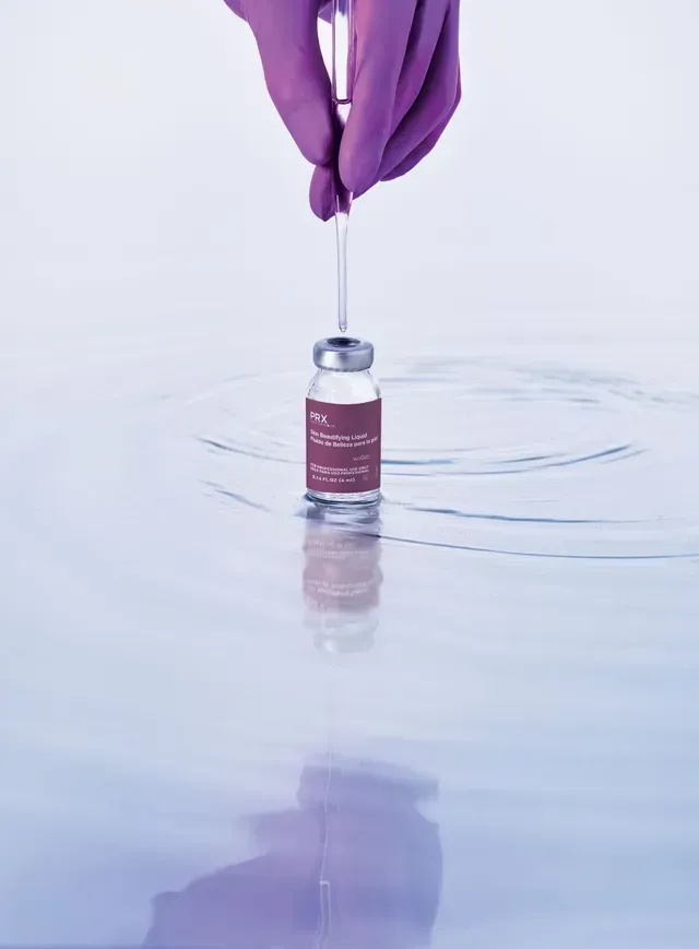 Purple-gloved hands hold a syringe above a small vaccine vial on a glossy surface.
