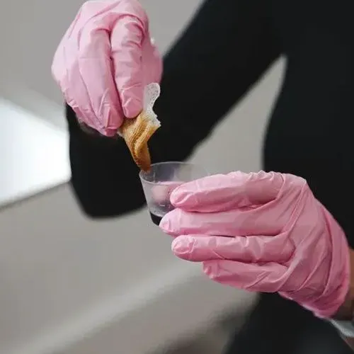 Gloved hands dip a pastry into a small cup of sauce or liquid.