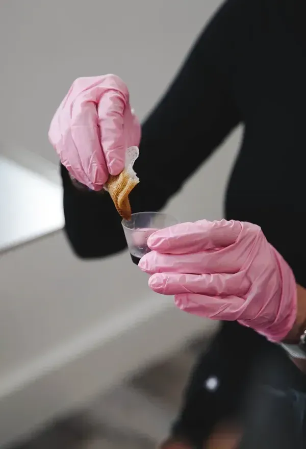 Pink-gloved hands holding a small pipette over a vial in a lab setting