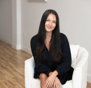 Woman with long dark hair sitting on a white chair in a bright room, smiling at the camera.