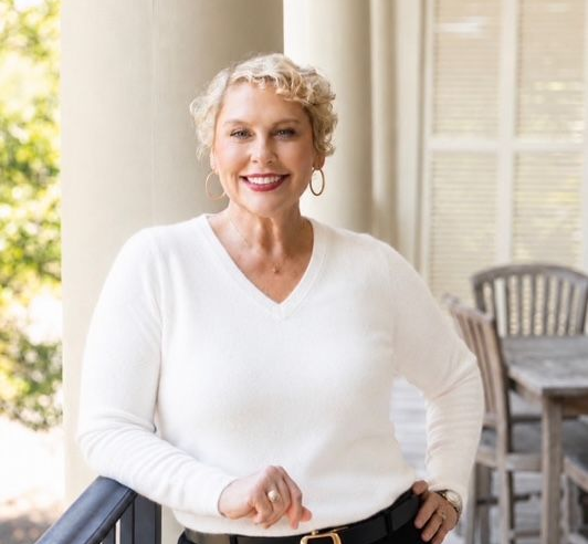 Smiling person in a white sweater standing on a porch with a chair and window behind them