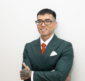 Man in a dark green suit with red tie and glasses, smiling with arms crossed against a white background