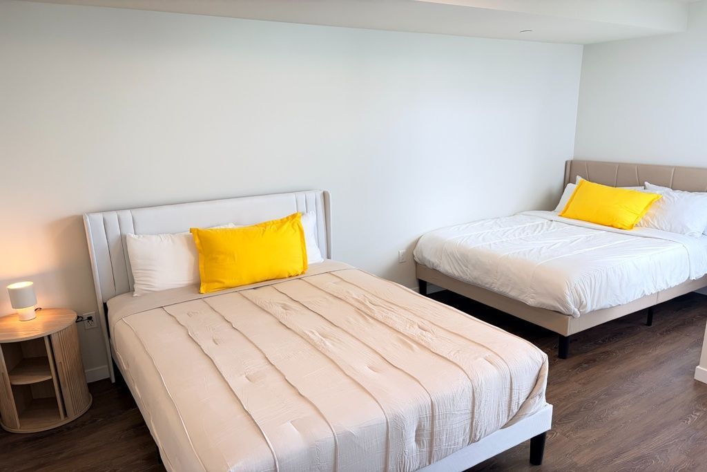 Bedroom with two neatly made beds, white bedding, and yellow accent pillows beside a small bedside lamp.