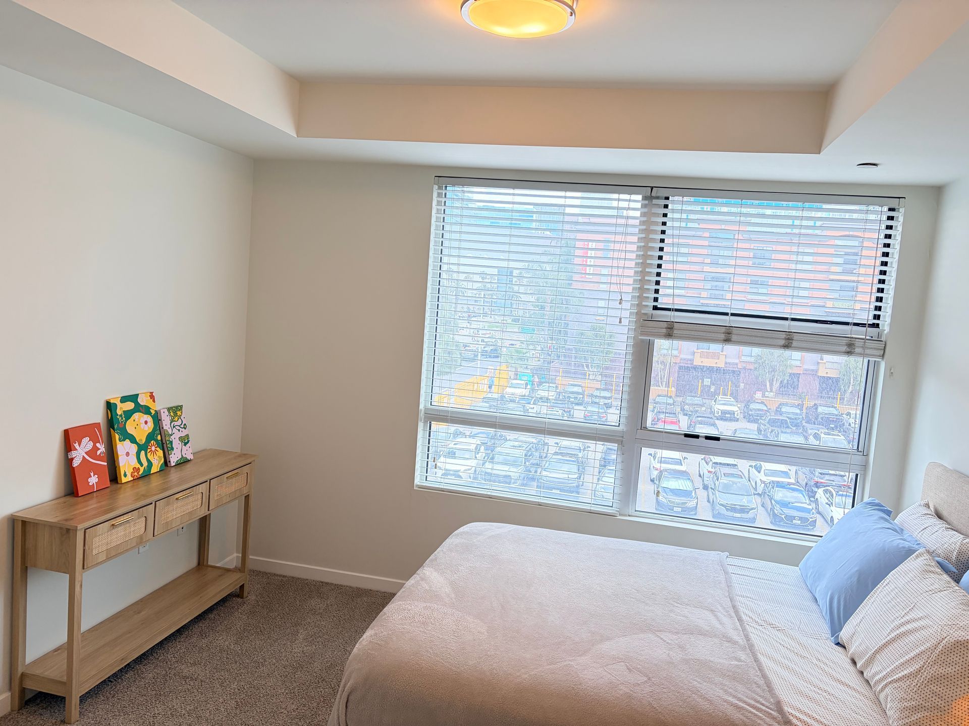 Bright bedroom with a bed, window blinds, beige carpet, and a small console table with decor.