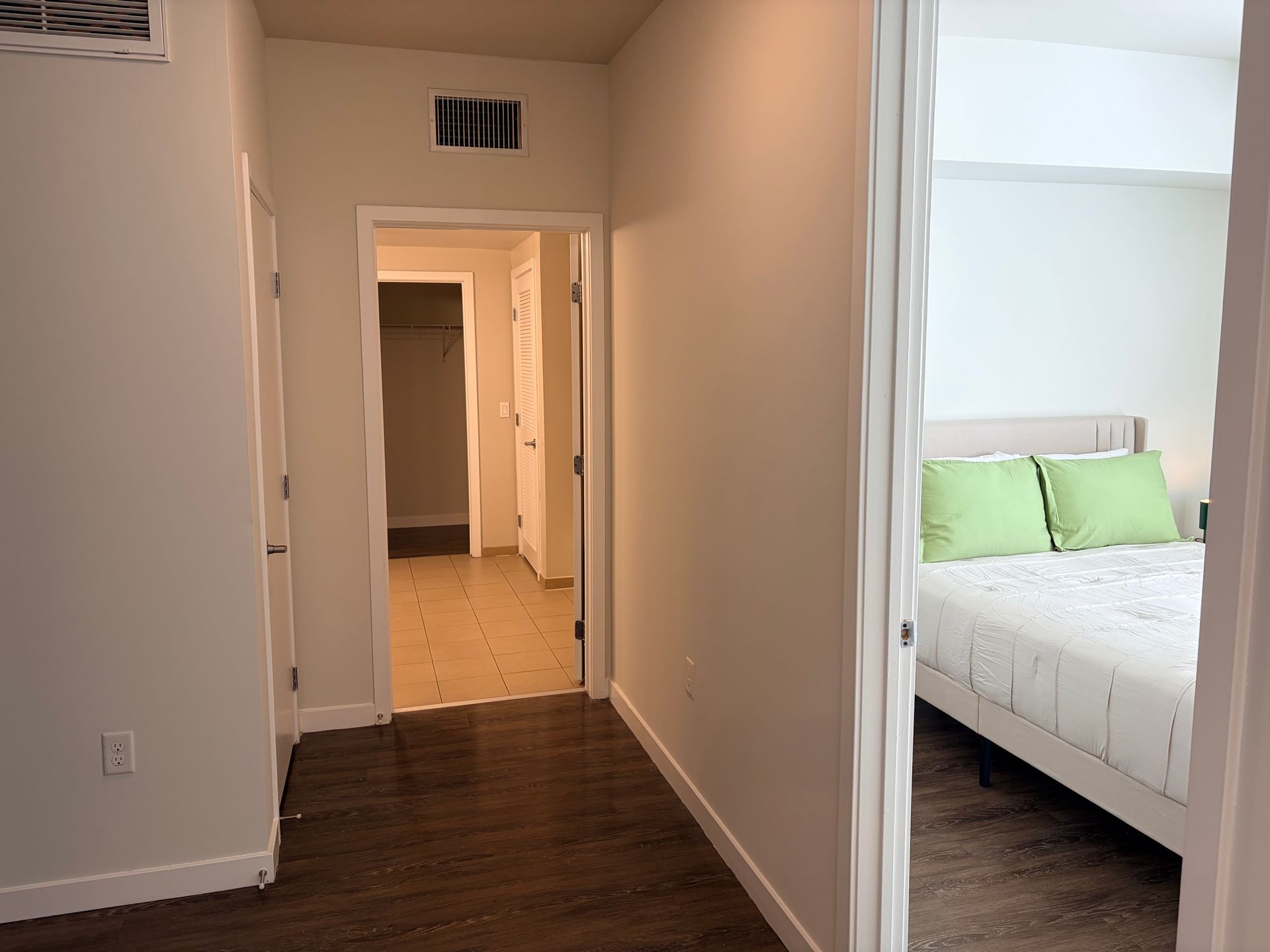 Hallway with white walls and dark wood floor, opening to a bedroom with white bedding and green pillows.