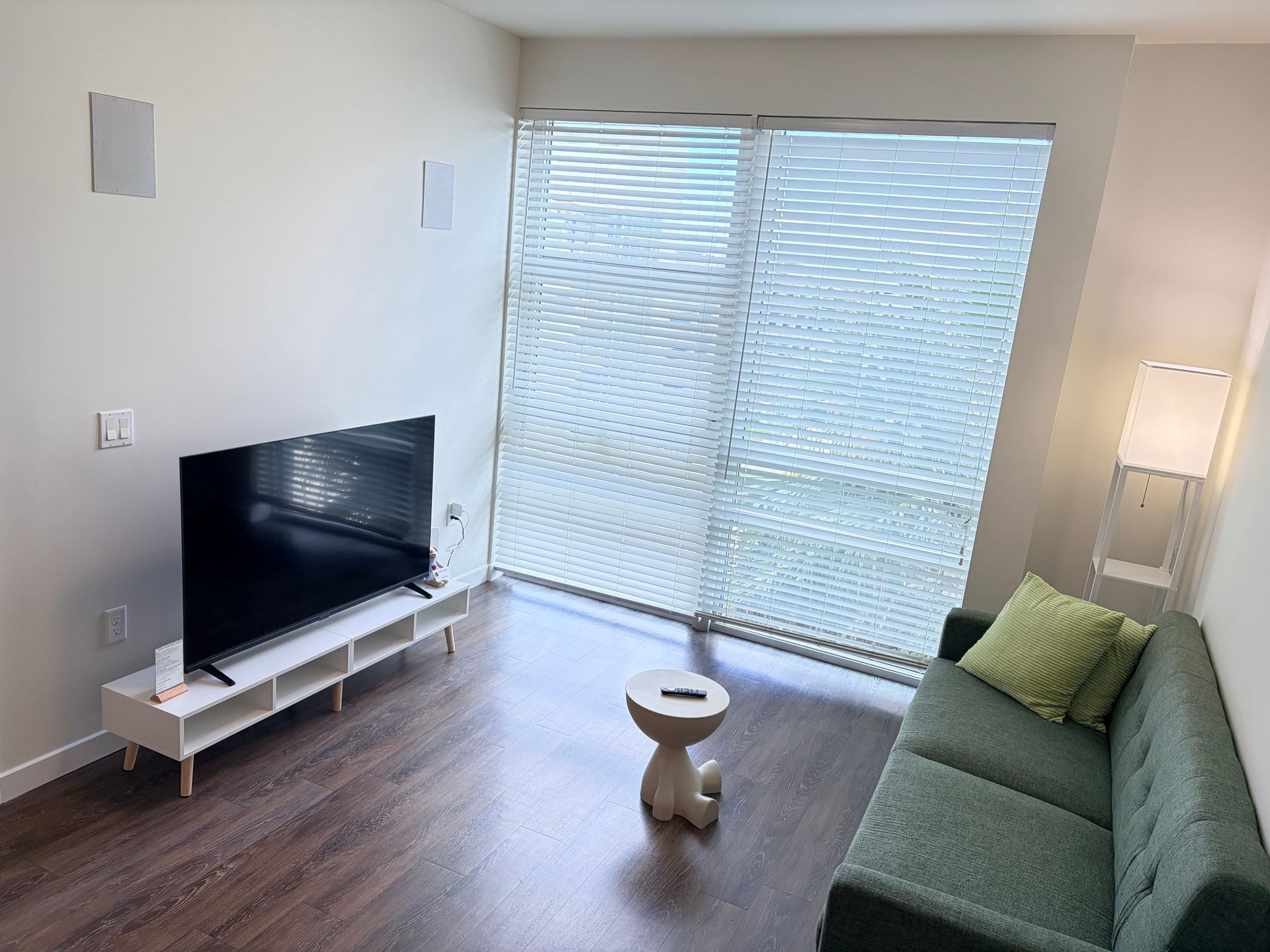 Bright living room with TV, green sofa, small table, and large window blinds.