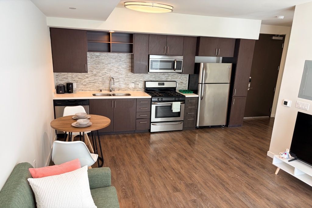 Modern apartment kitchen and living area with stainless steel appliances, wood floors, and green sofa corner
