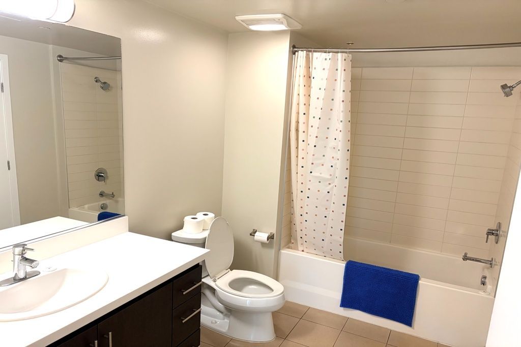 Bright bathroom with sink, mirror, toilet, and bathtub with beige shower curtain and blue bath mat
