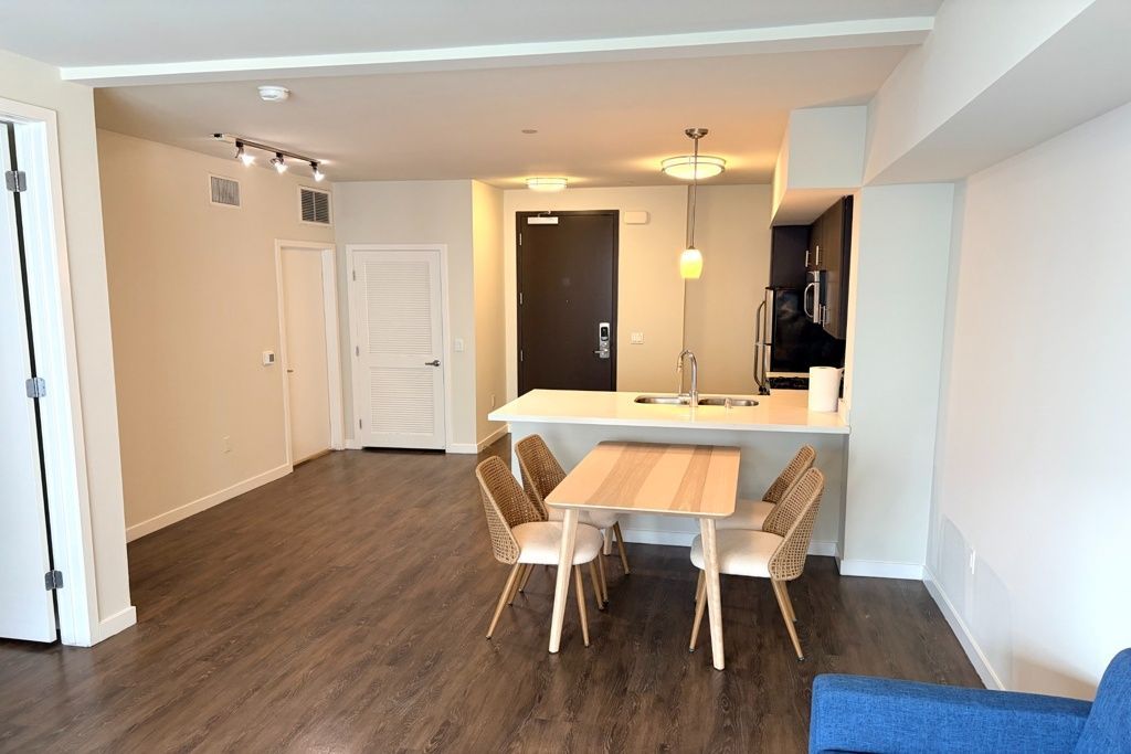 Open living-dining room with wood floors, white walls, a table and chairs, and a kitchen pass-through.