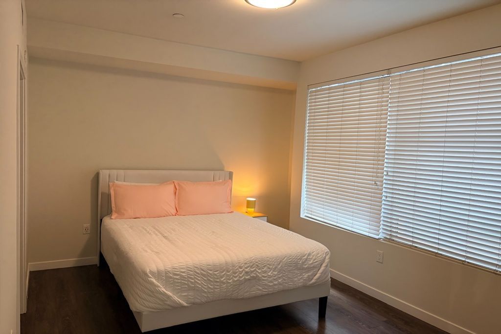 Bedroom with bed, pink pillows, bedside lamp, and large window with blinds.