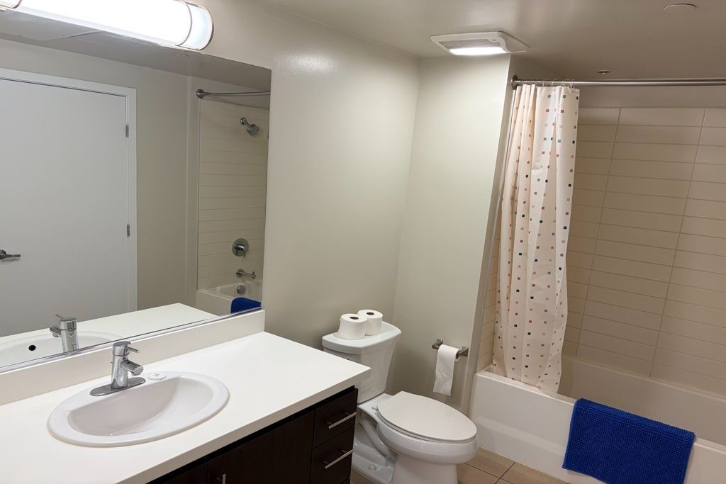 Bright bathroom with sink, mirror, toilet, tub, and beige shower curtain with blue towel.