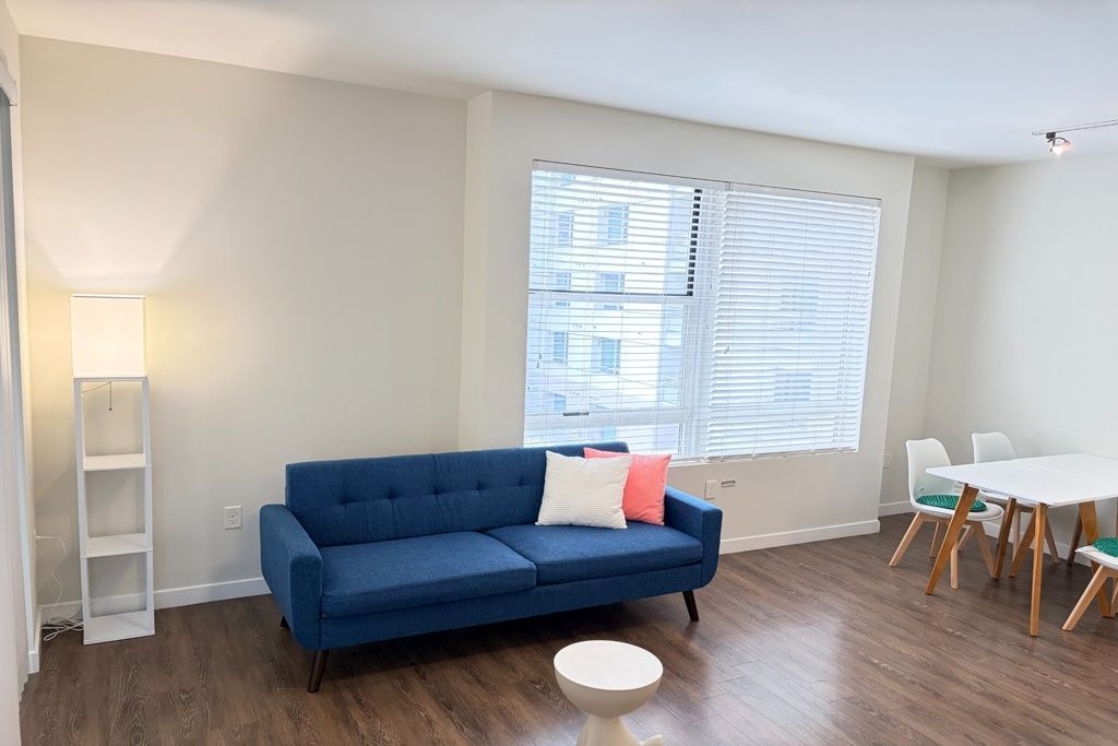 Bright living room with blue sofa, white floor lamp, large window, and small dining table with chairs.